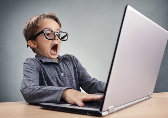 Wearing glasses, a surprised boy works on a laptop. 