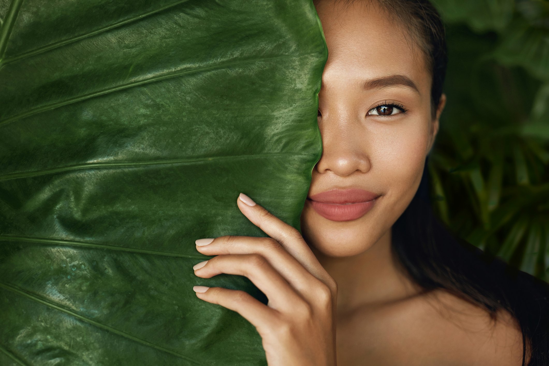 A young womanin makeup peers out from behind a large green palm leaf. 