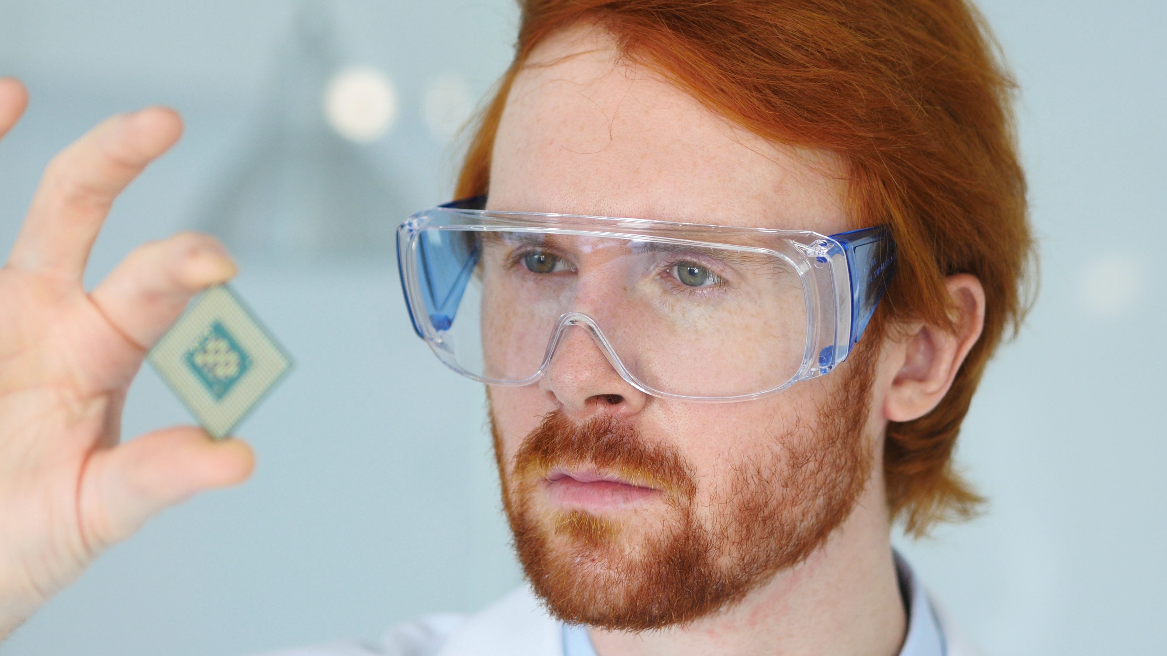 A technician wearing safety goggles holds up an integrated circuit for a close inspection.