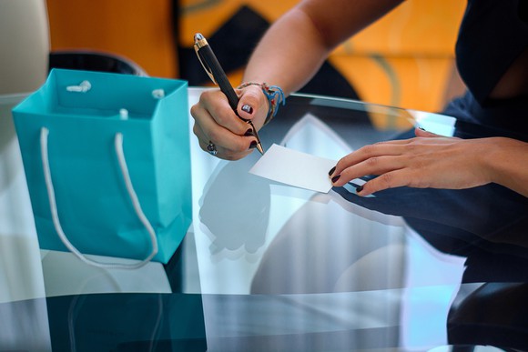 A woman writes a note on a glass table with a small blue bag.