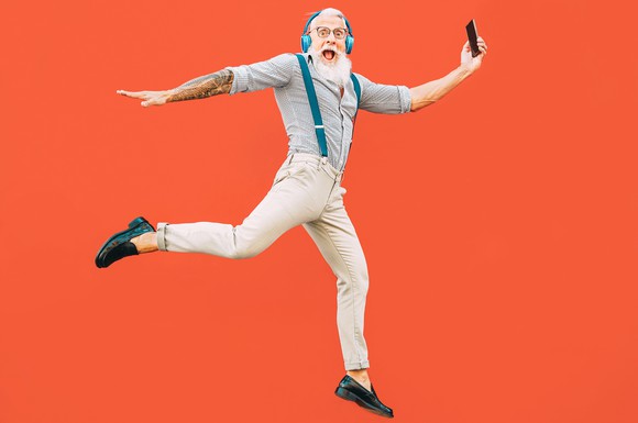 An oder man with a beard in suspenders leaps while wearing headphones and holding a cell phone. 