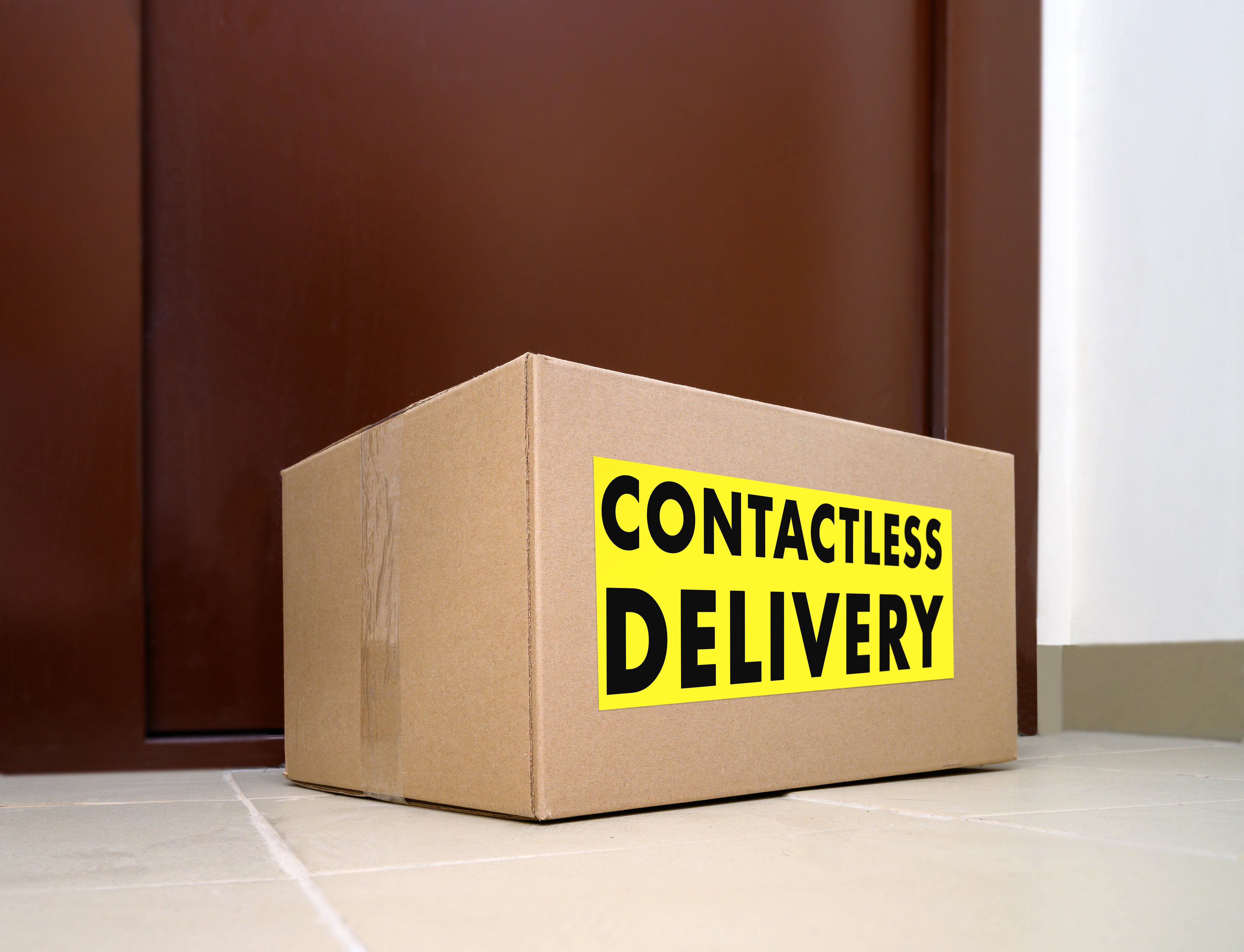 A cardboard box with 
a label that says "contactless delivery" sits outside a front door.