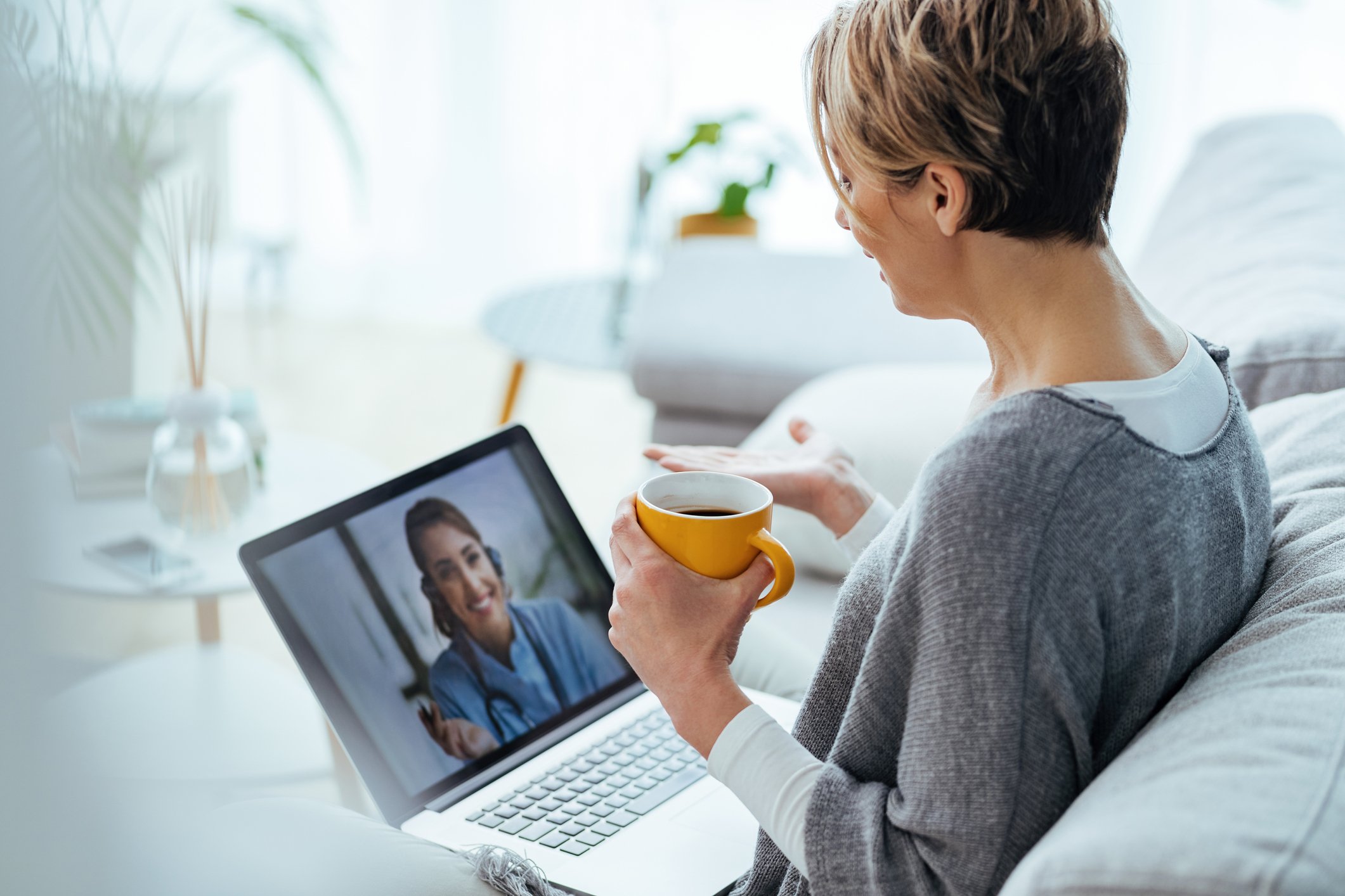 A woman sits on a white couch holding a mug and talking to another woman through a laptop video chat. 