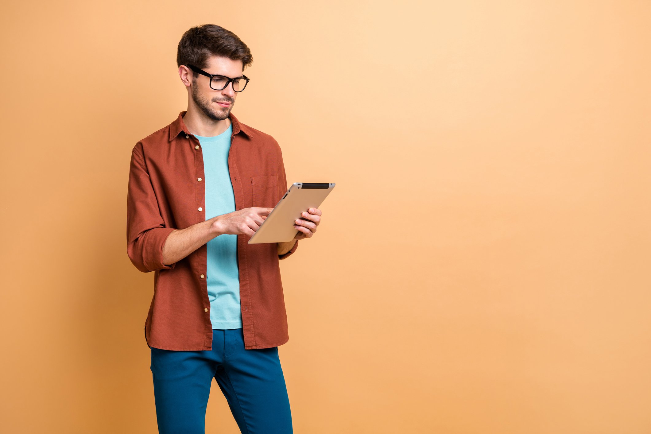 A young man in casual wear and glasses holds and works an electronic tablet. 