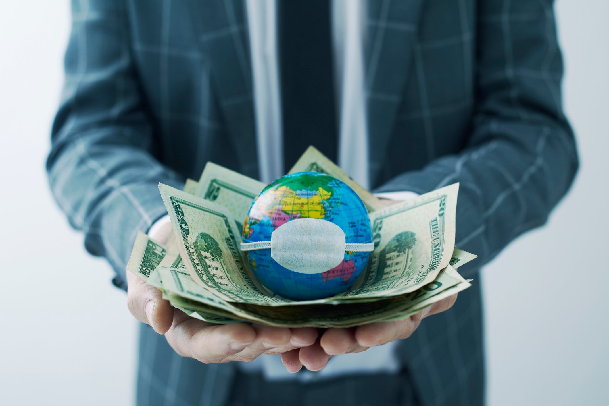 A businessman holds a pile of dollars and a globe wearing a mask.
