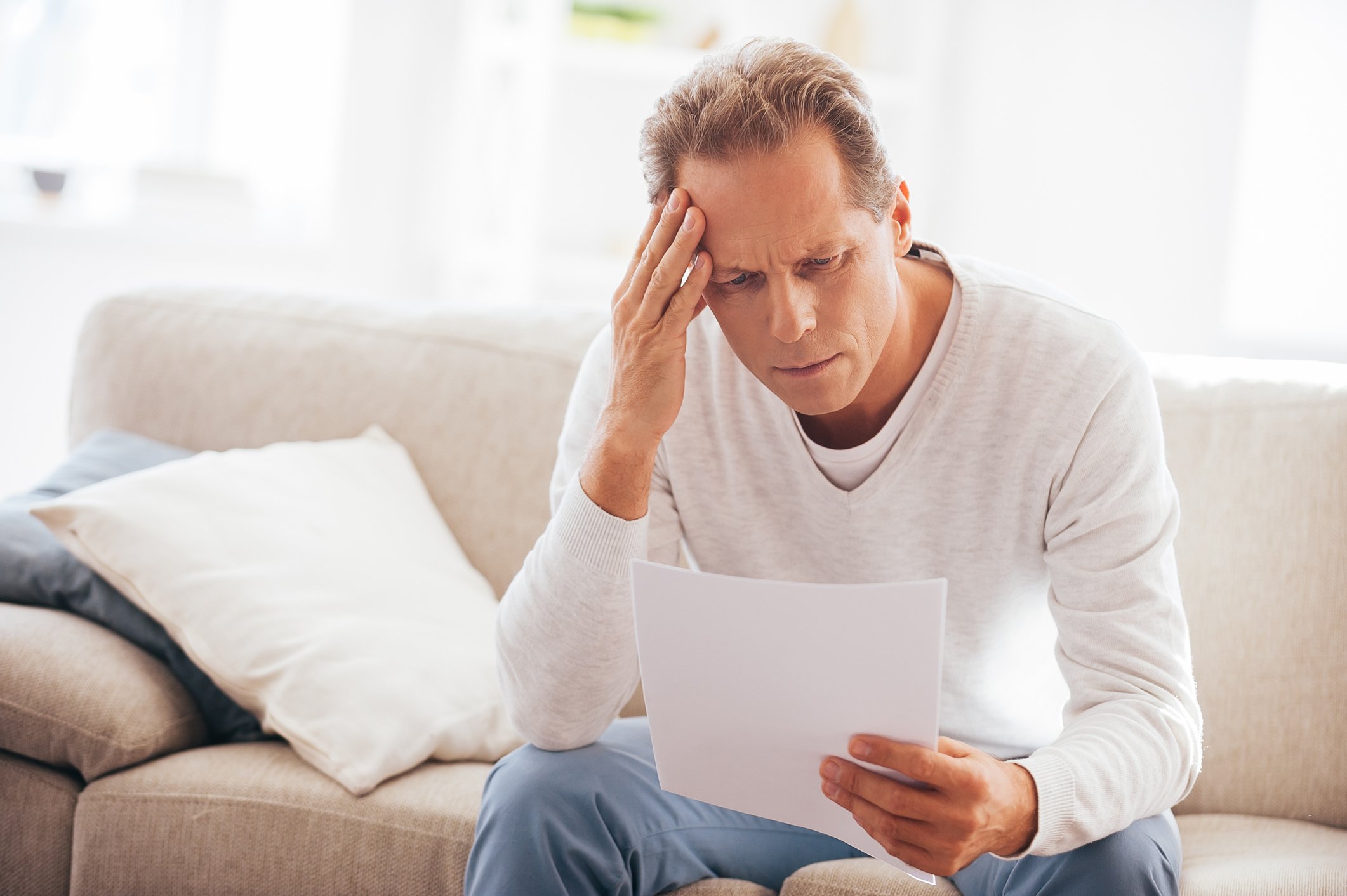Man sitting on couch looking at document with pained expression