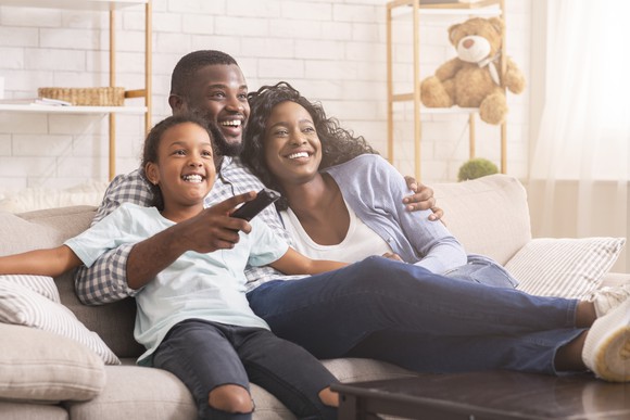 A young family smiles while sitting on the couch and watching a movie.