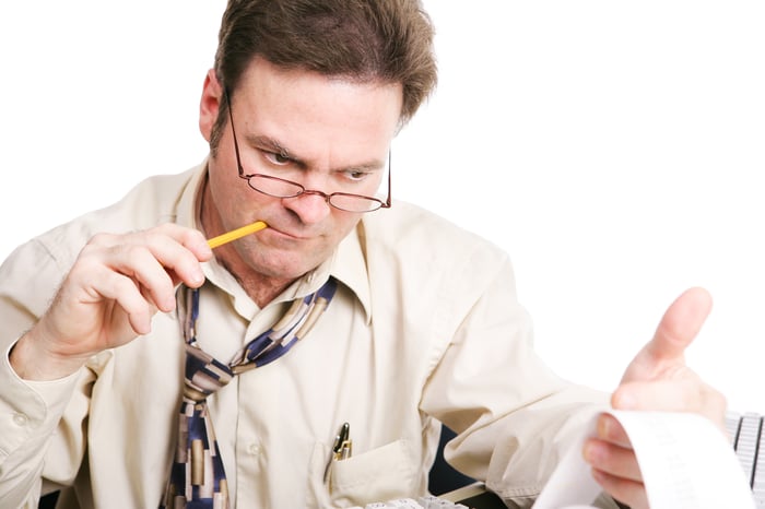 A man chewing on a pencil while closely analyzing figures from his printing calculator. 