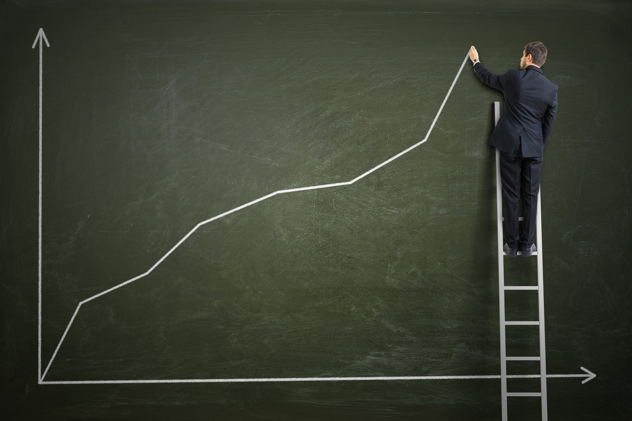 Man on a ladder drawing an upward pointing graph on a board. 