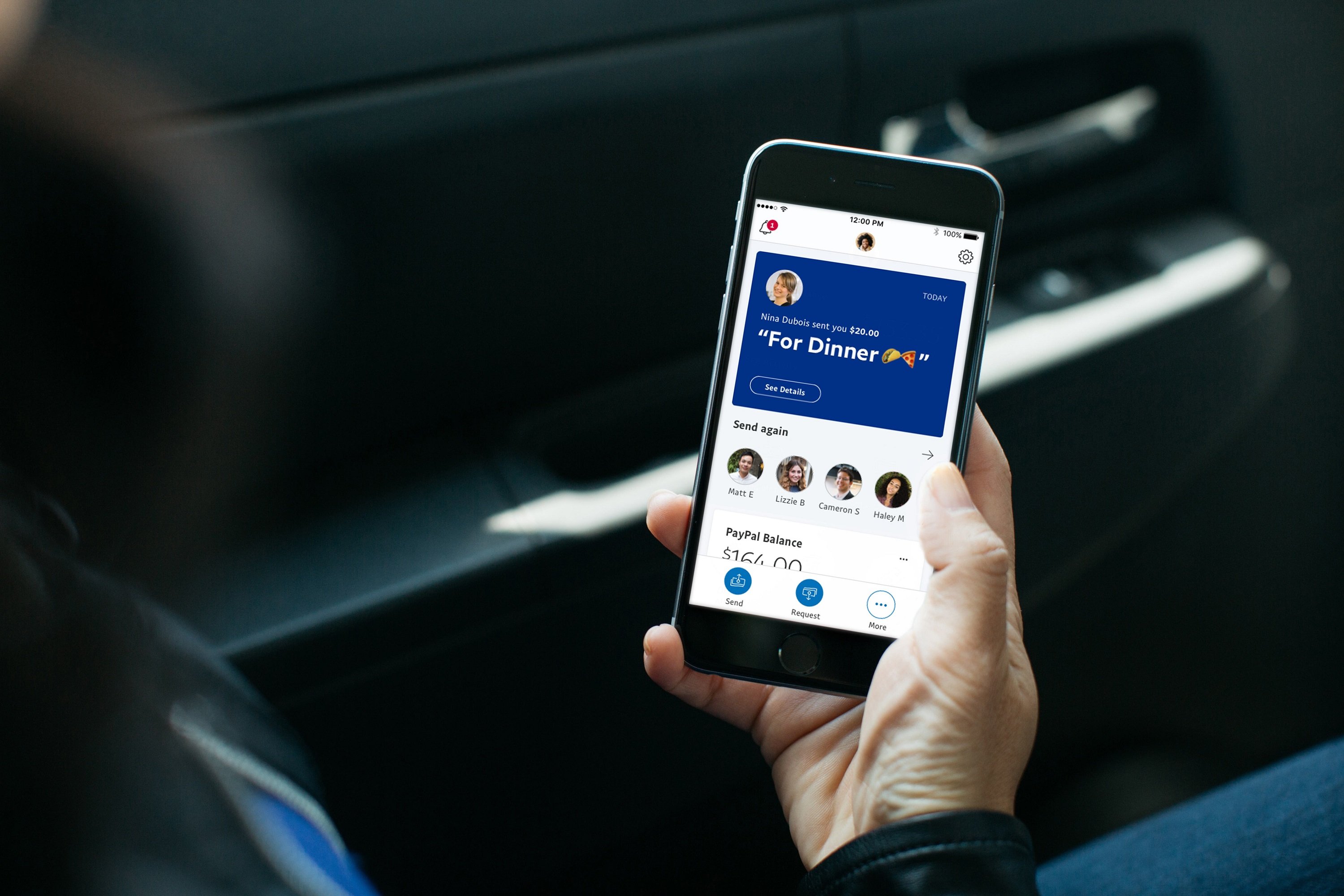A person sitting in a car holding a smartphone looking at the PayPal app homescreen.
