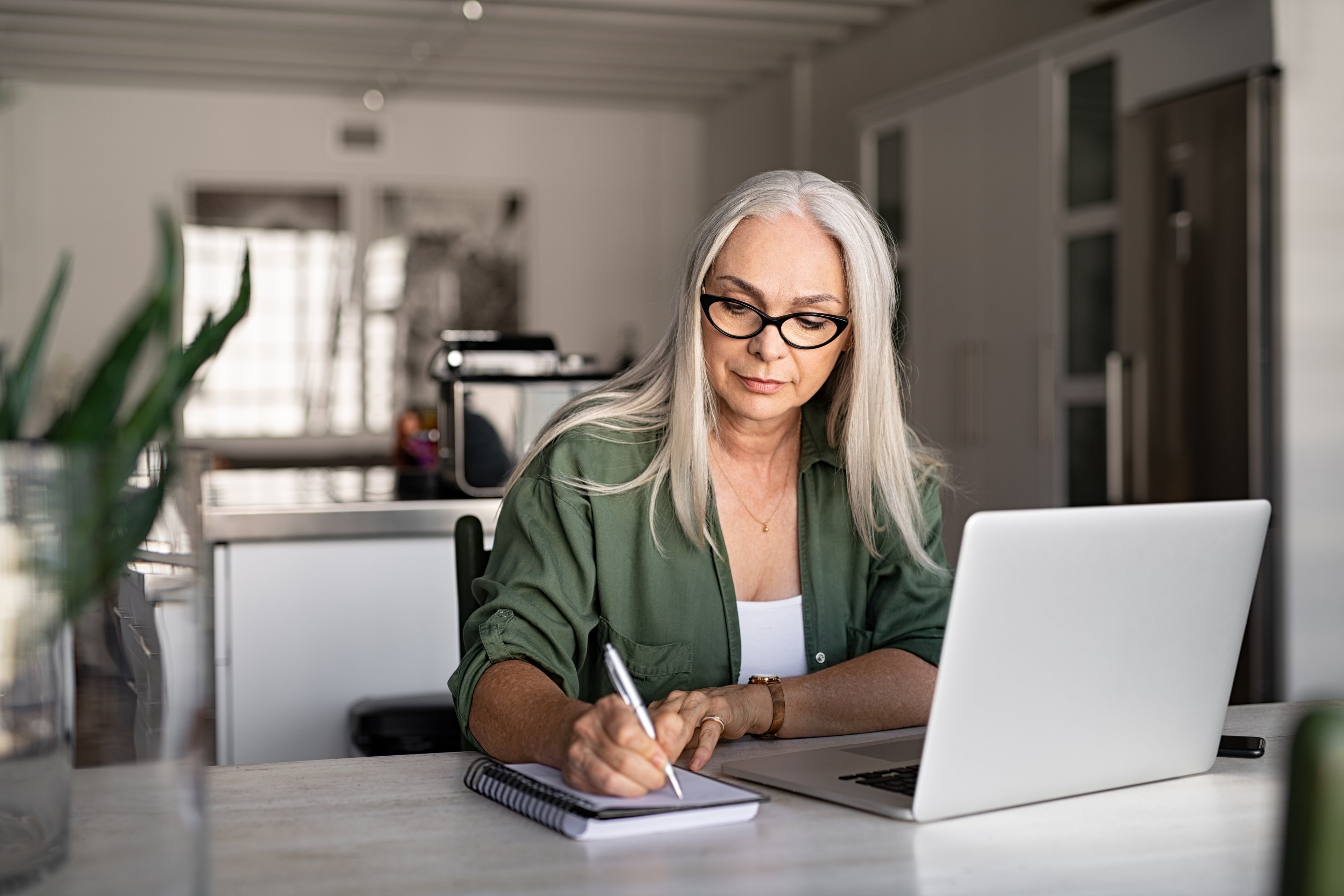 Woman sitting at computer, writing on notepad.