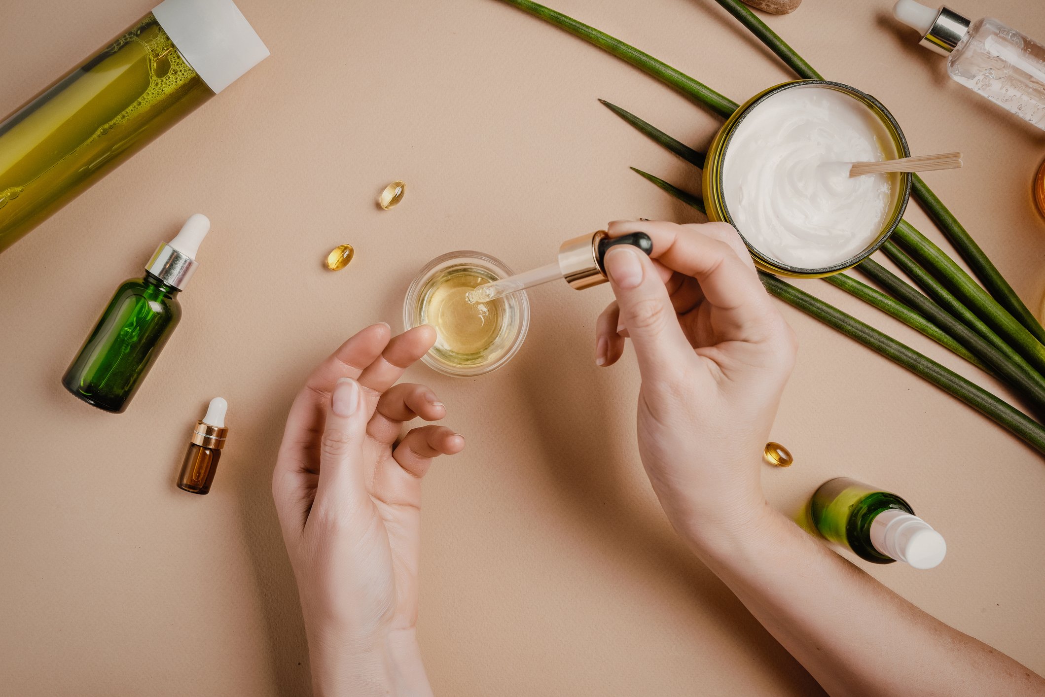 A woman's hands are surrounded by bottles, jars, and plants, suggesting natural products, as she fills a dropper from one jar.
