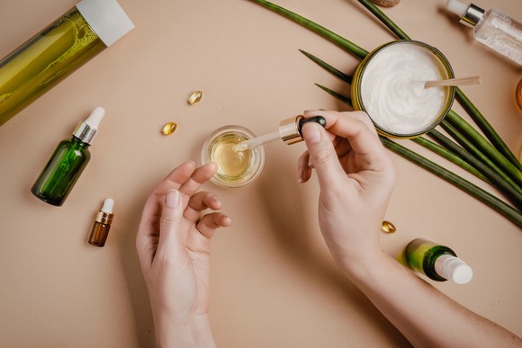 A woman's hands are surrounded by bottles, jars, and plants, suggesting natural products, as she fills a dropper from one jar.