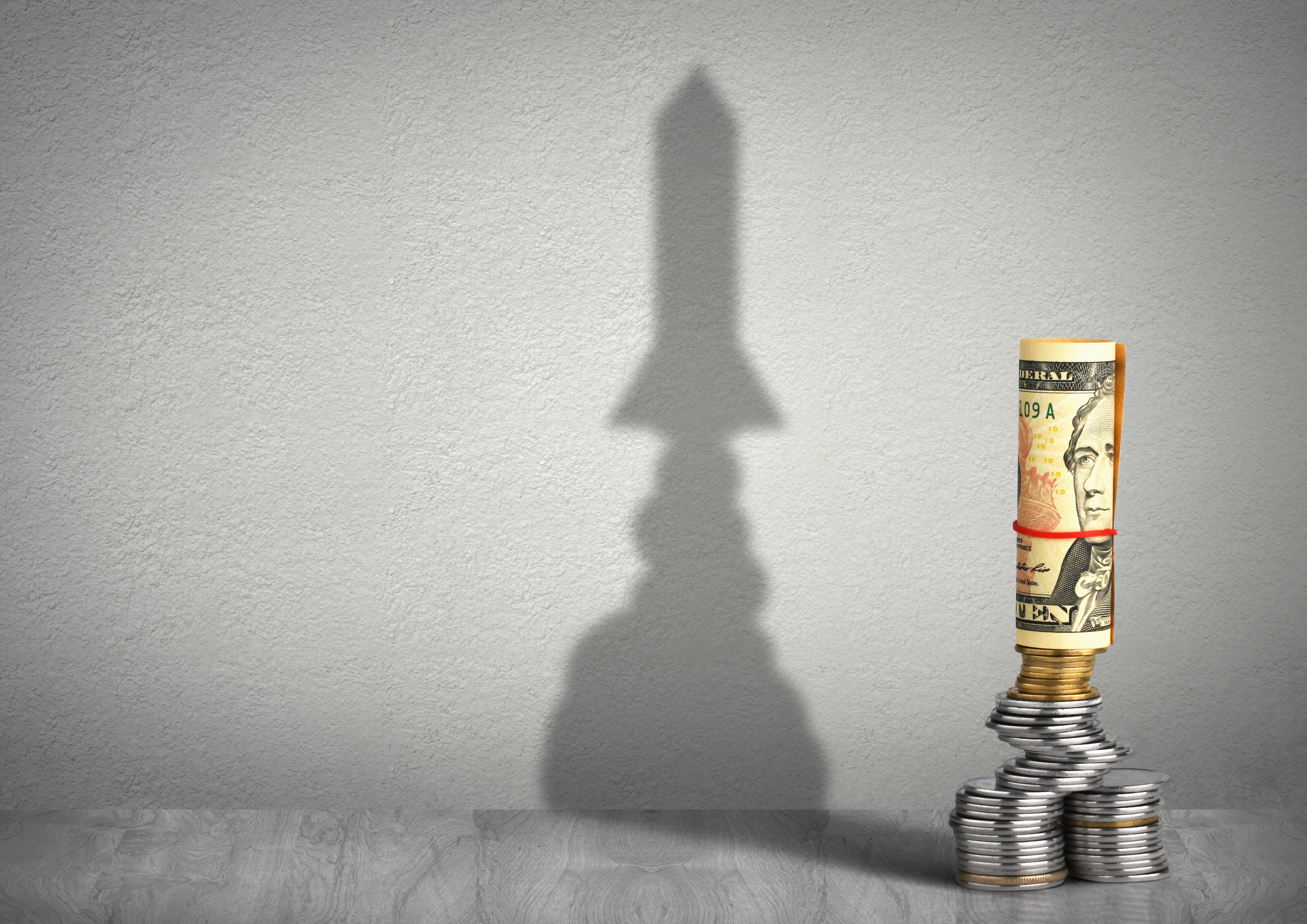A roll of hundred-dollar bills and quarter coins cast a shadow in the shape of a rocket taking off.