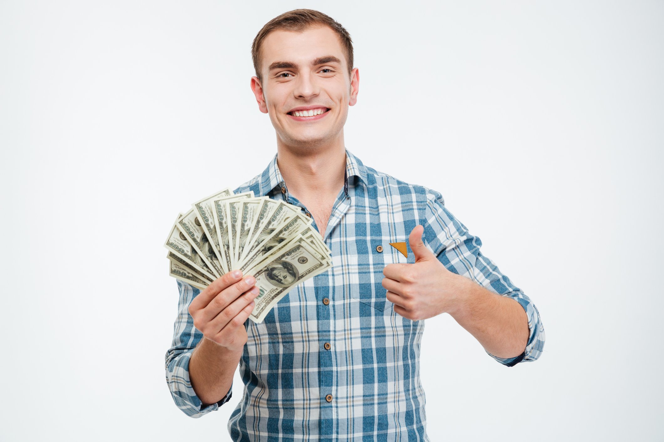 A young smiling white man in a blue plaid shirt holds a bunch of hundred dollar bills fanned out in one hand while giving a thumbs-up sign with the other. 