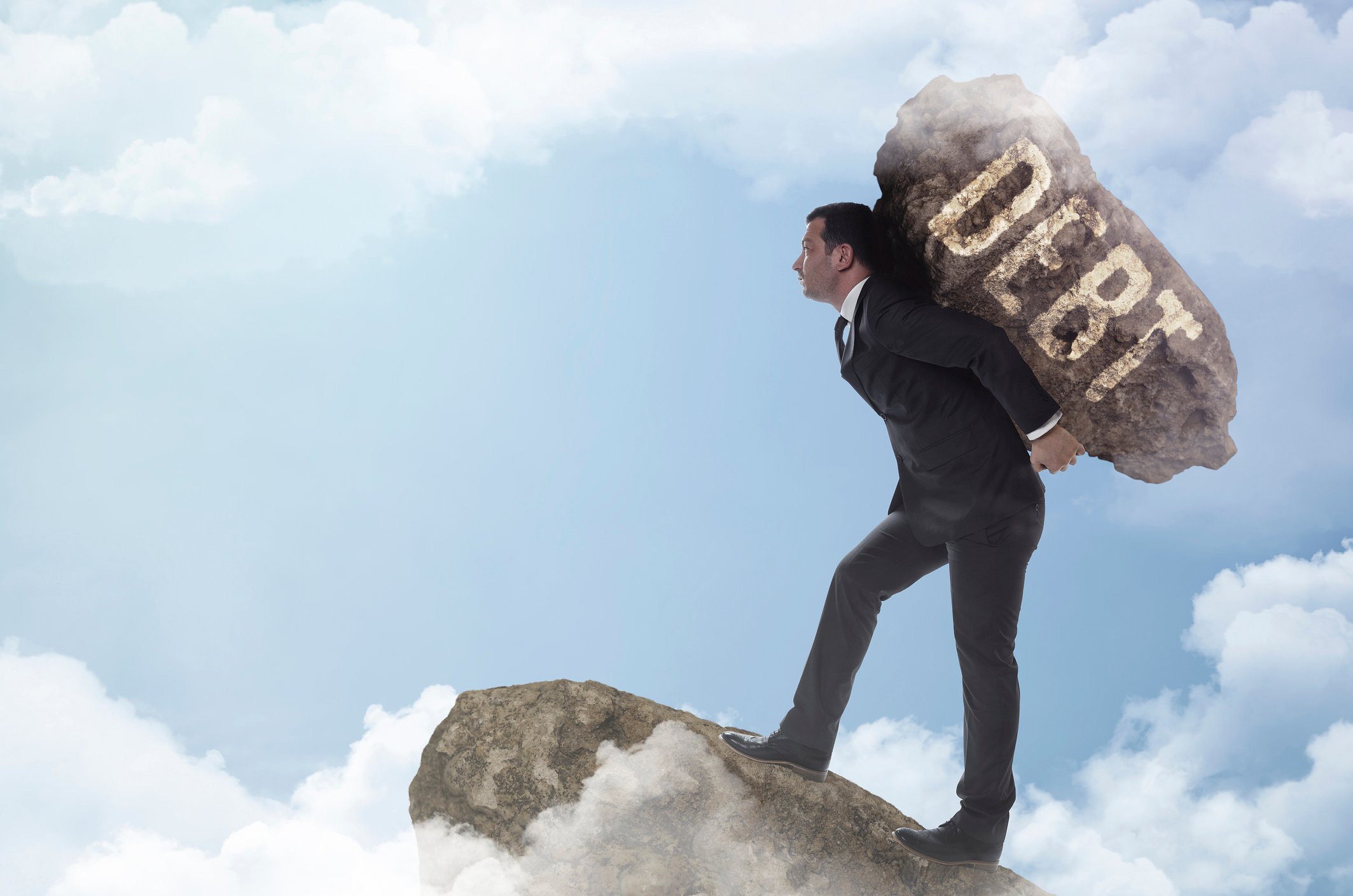 Man in a suit carrying a large rock labeled debt on his back up a mountain 