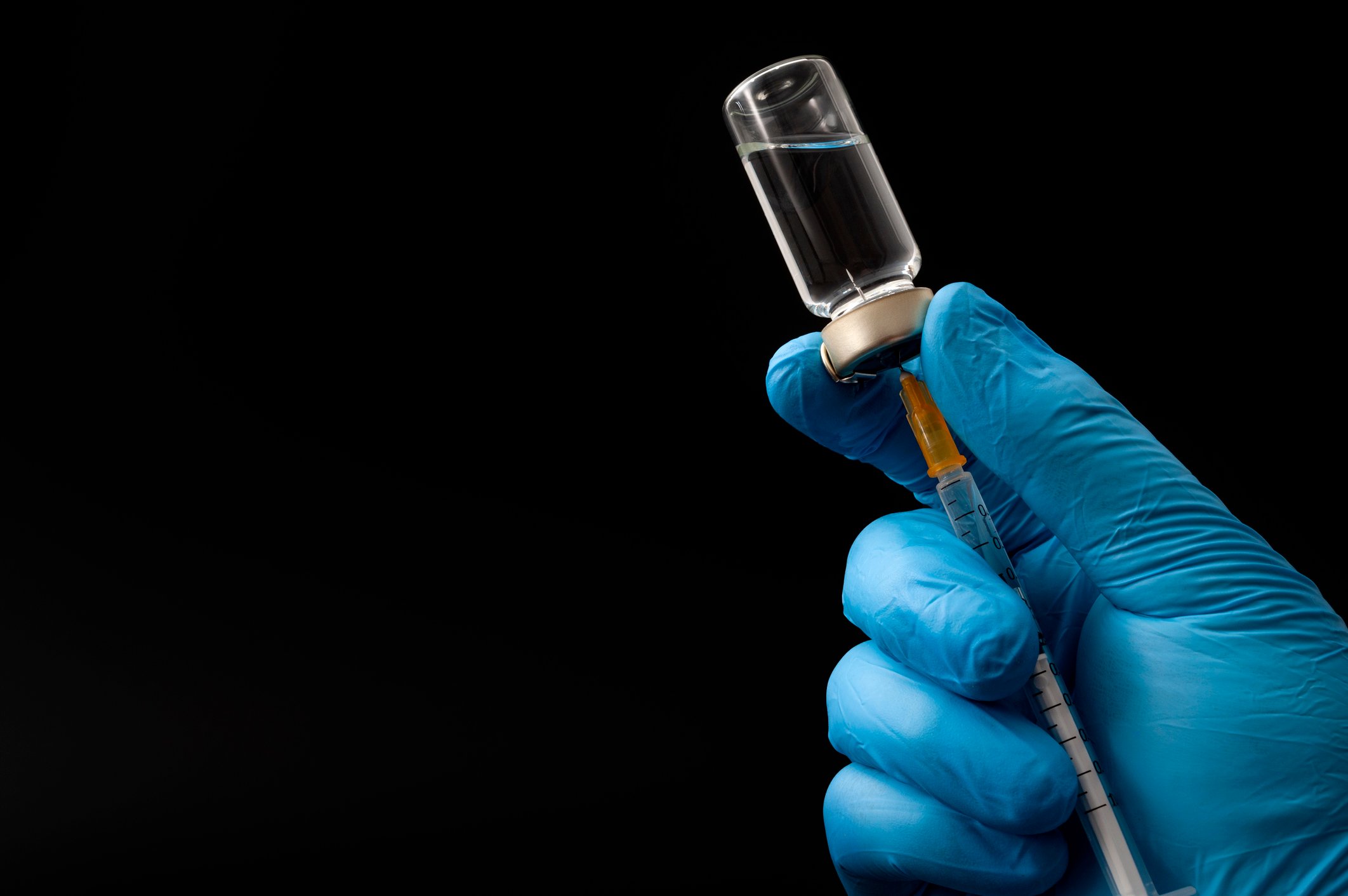 A hand in a blue nitrile exam glove fills a syringe with clear liquid from glass vial. 
