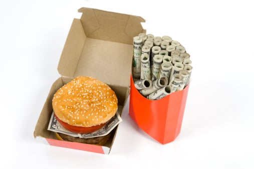 A burger with a five-dollar bill instead of lettuce in a cardboard carton next to a paper French fry box stuffed with tightly rolled dollar bills. 