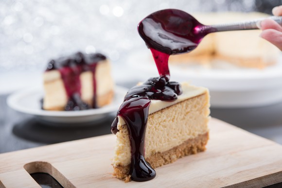Slice of cheesecake on a cutting board with someone drizzling  a topping over the dessert. 