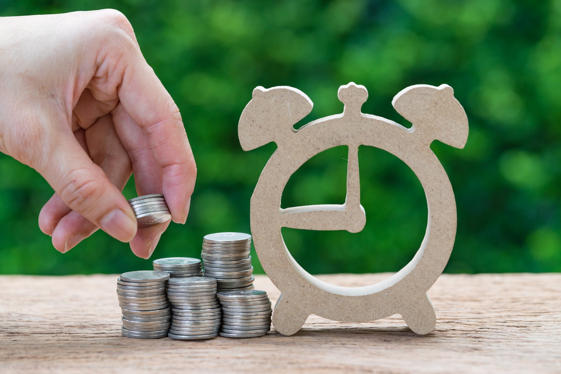 A hand stacks coins near a wooden cutout of an alarm clock.