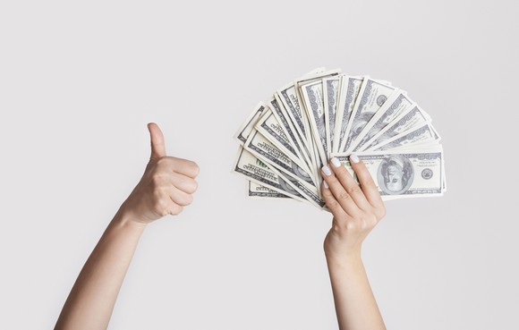 Thumb up with a hand holding $100 bills fanned out