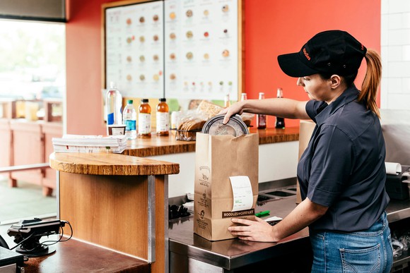 A Noodles & Company employee places food in a bag for a to-go order.