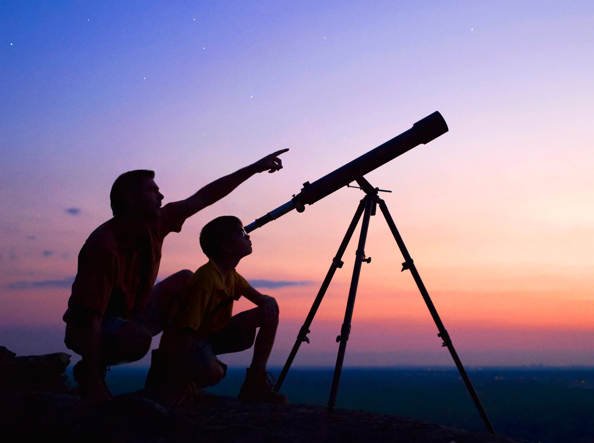 A father and son look through a telescope at sunset. 