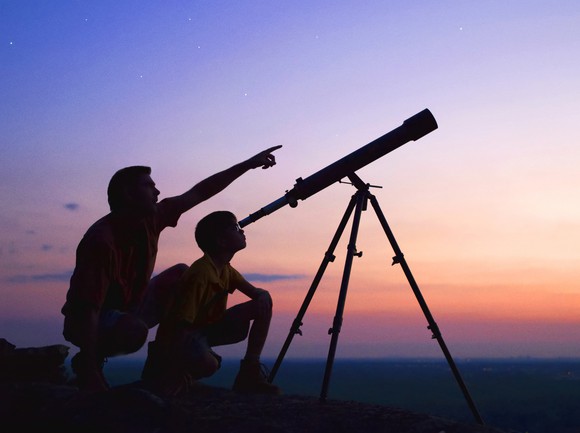 A father and son look through a telescope at sunset. 