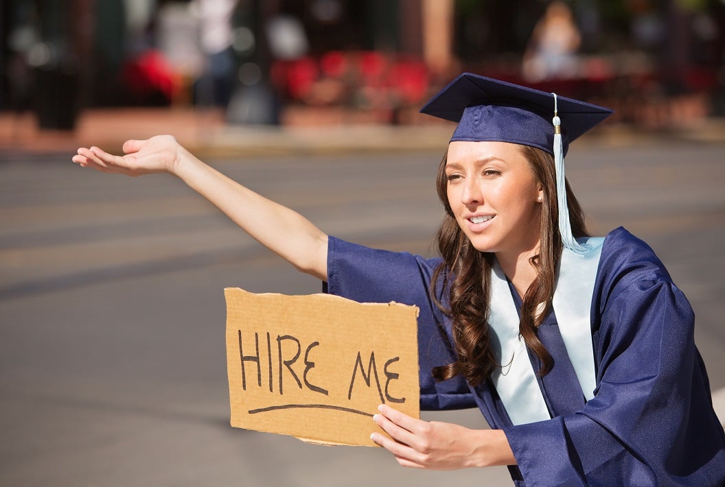 A woman wearing a graduation gown and cap holds her hand out with a sign saying Hire Me.