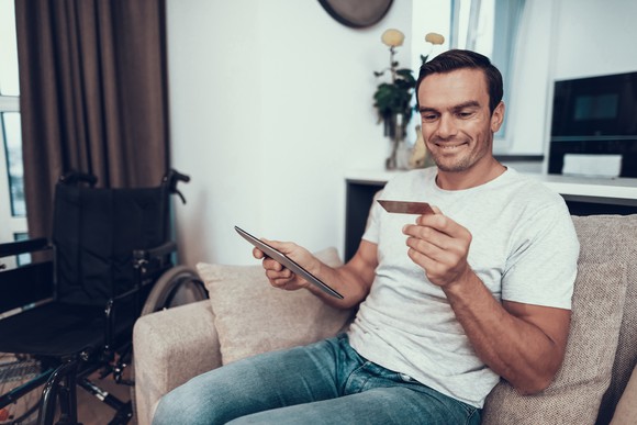 A man sits on his couch next to a wheelchair and shops online.