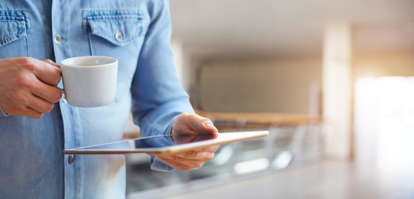 A man drinks coffee while reading his tablet.