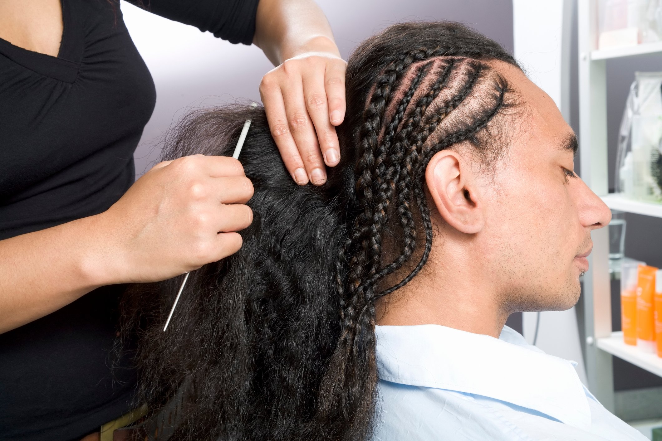 Woman styling a man's hair in multiple braids at a salon.