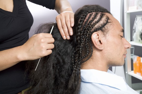 Woman styling a man's hair in multiple braids at a salon.