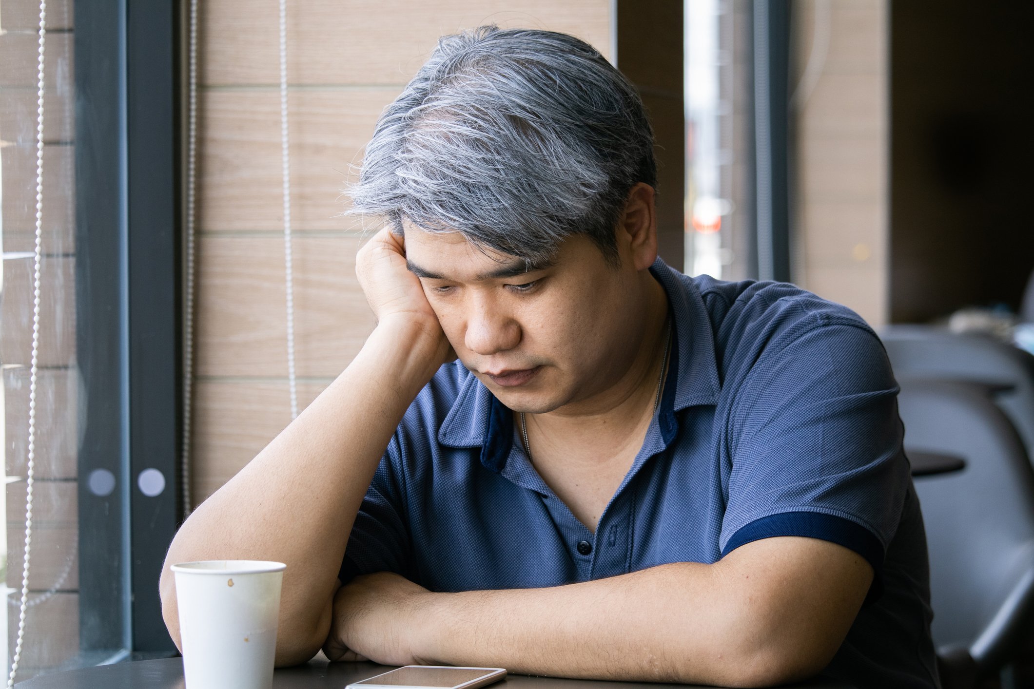 A sad man hangs his head sitting at a cafe table with a paper cup of coffee. 