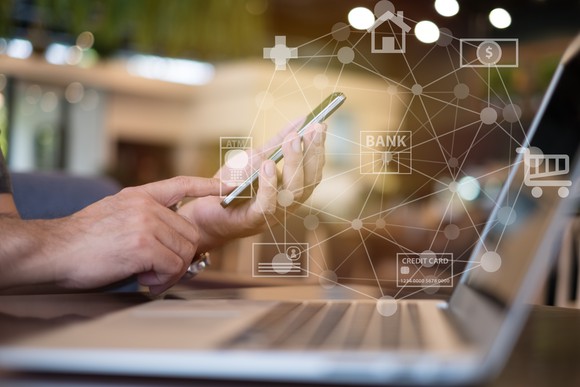 hands hold a smartphone near a laptop computer in the foreground as digital banking icons overlay the image.