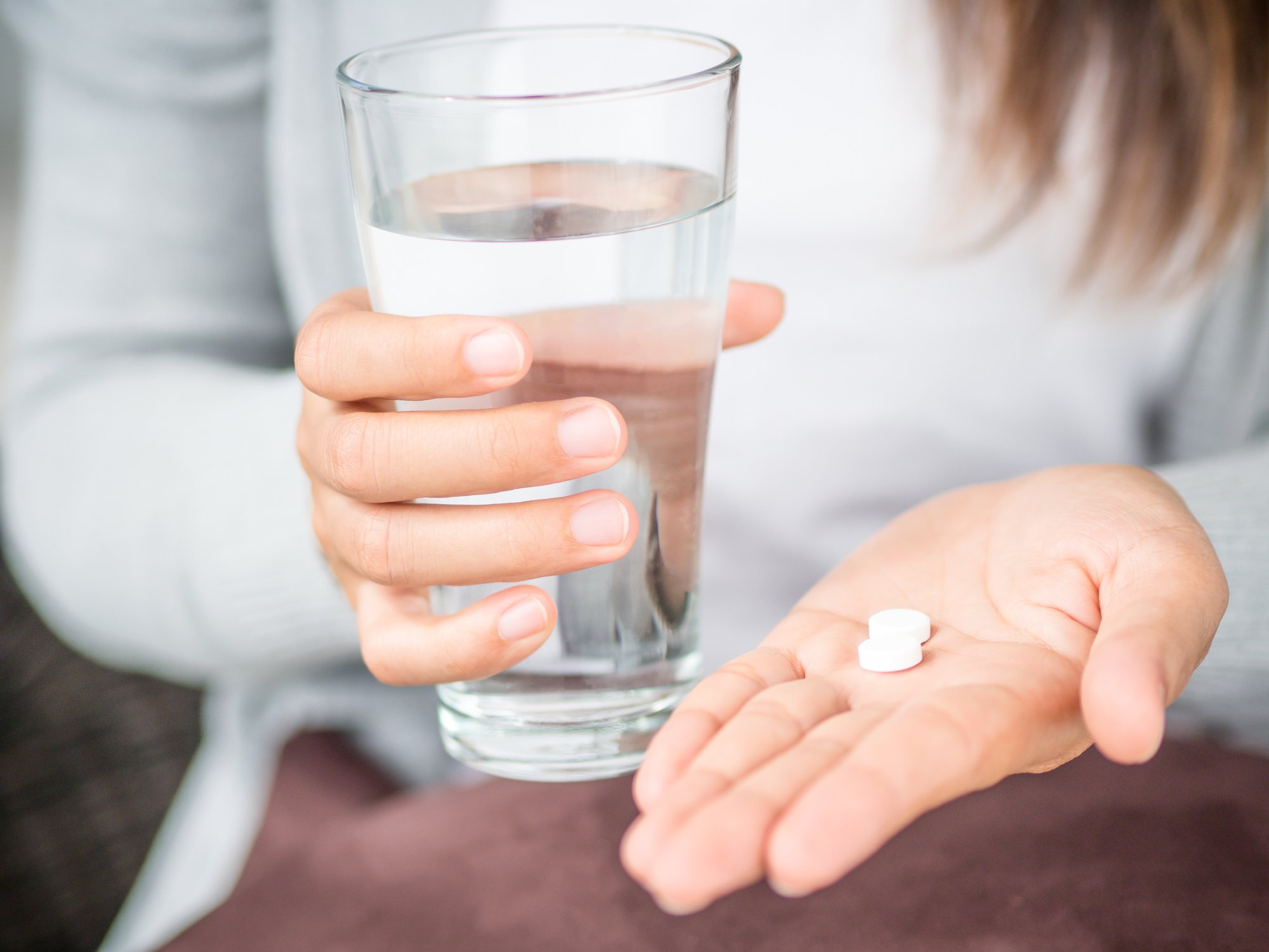 A woman holds a glass of water and shows two pills in her other hand.
