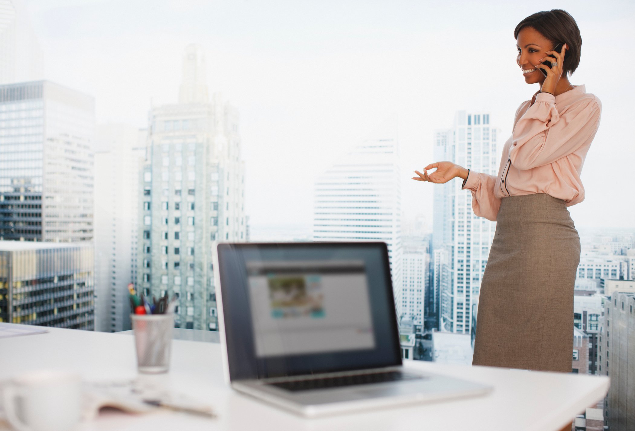 A smiling businesswoman talks on her phone in a home office with big-city views.