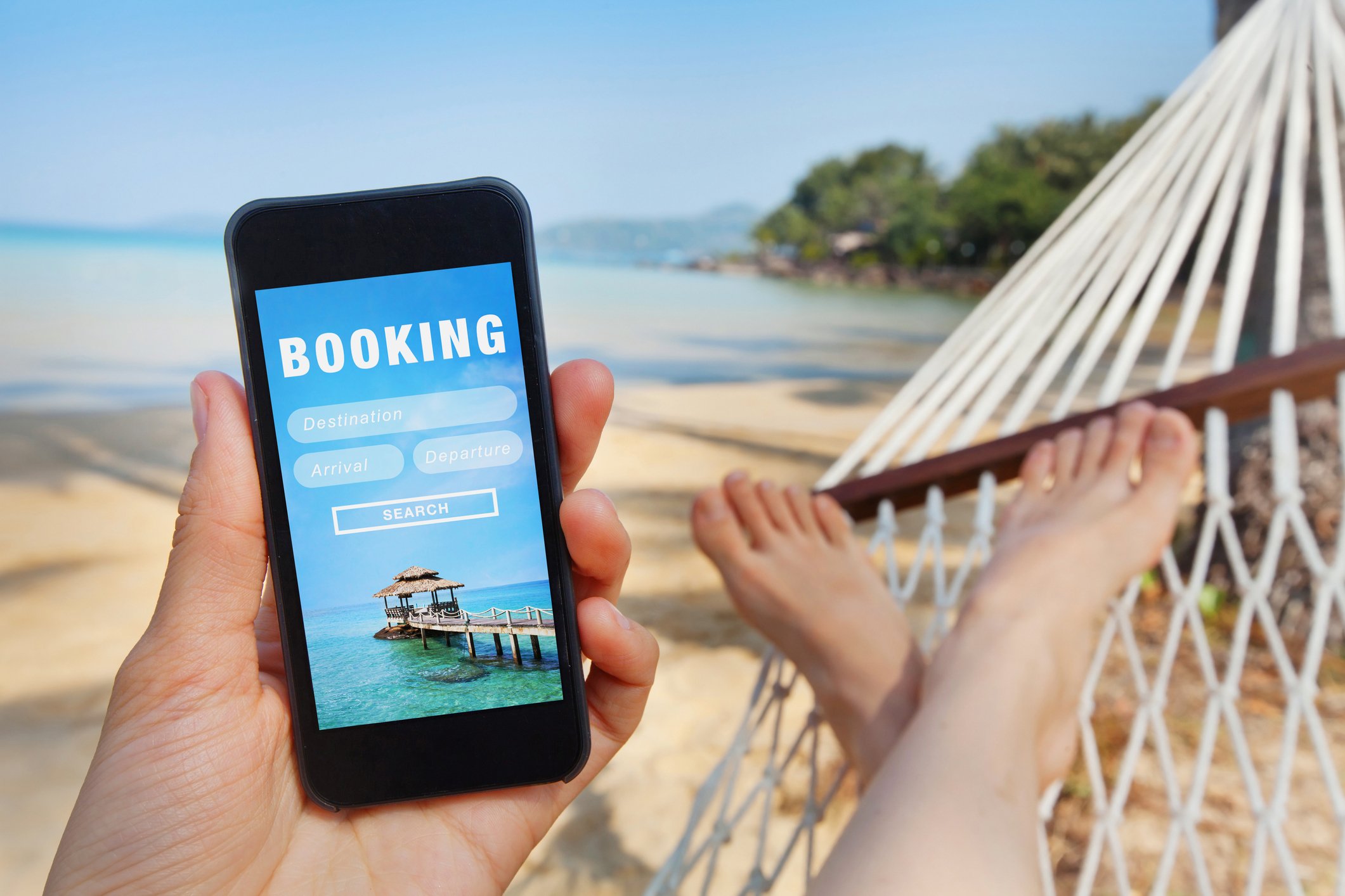 A person lies in a hammock, looking at a travel booking site on their phone.