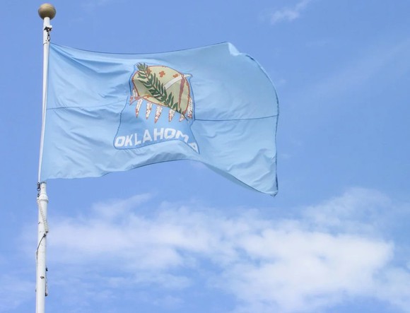 Oklahoma state flag flying on a flagpole against a blue sky with a few clouds.