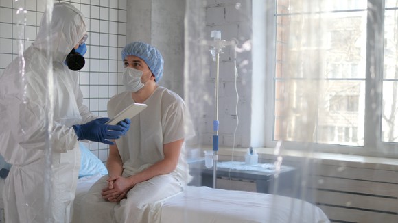 Doctor in protective equipment talking to face-masked patient seated on bed.
