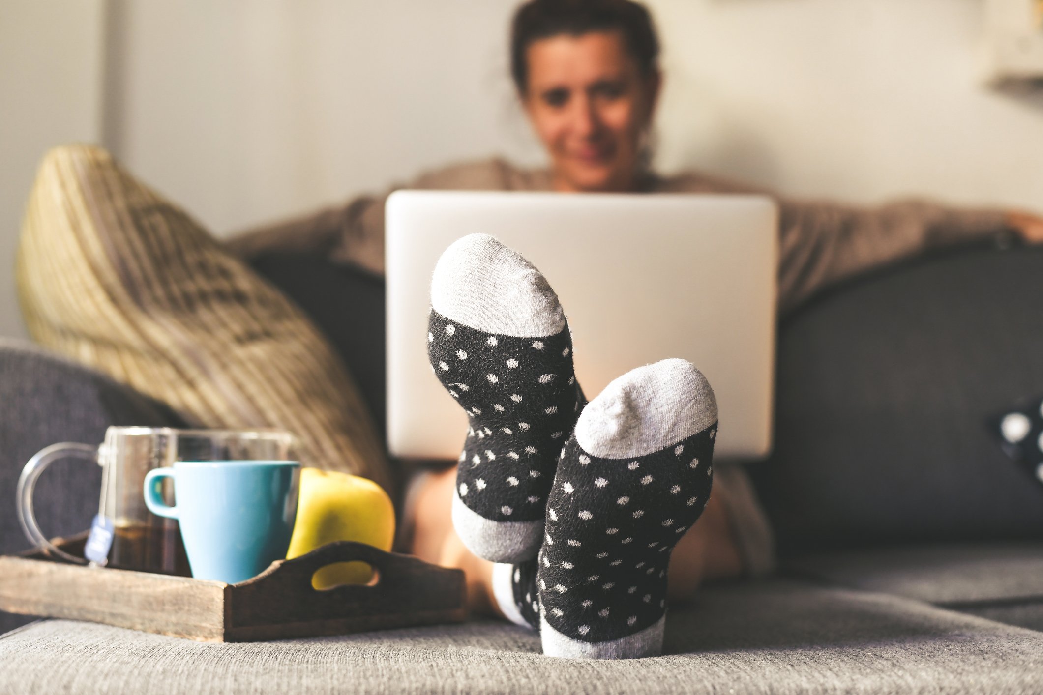 A woman on her couch with her laptop and several coffee mugs on a tray on her ottoman. 