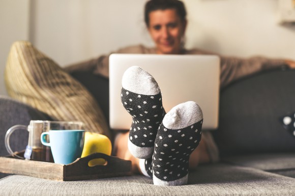 A woman on her couch with her laptop and several coffee mugs on a tray on her ottoman. 
