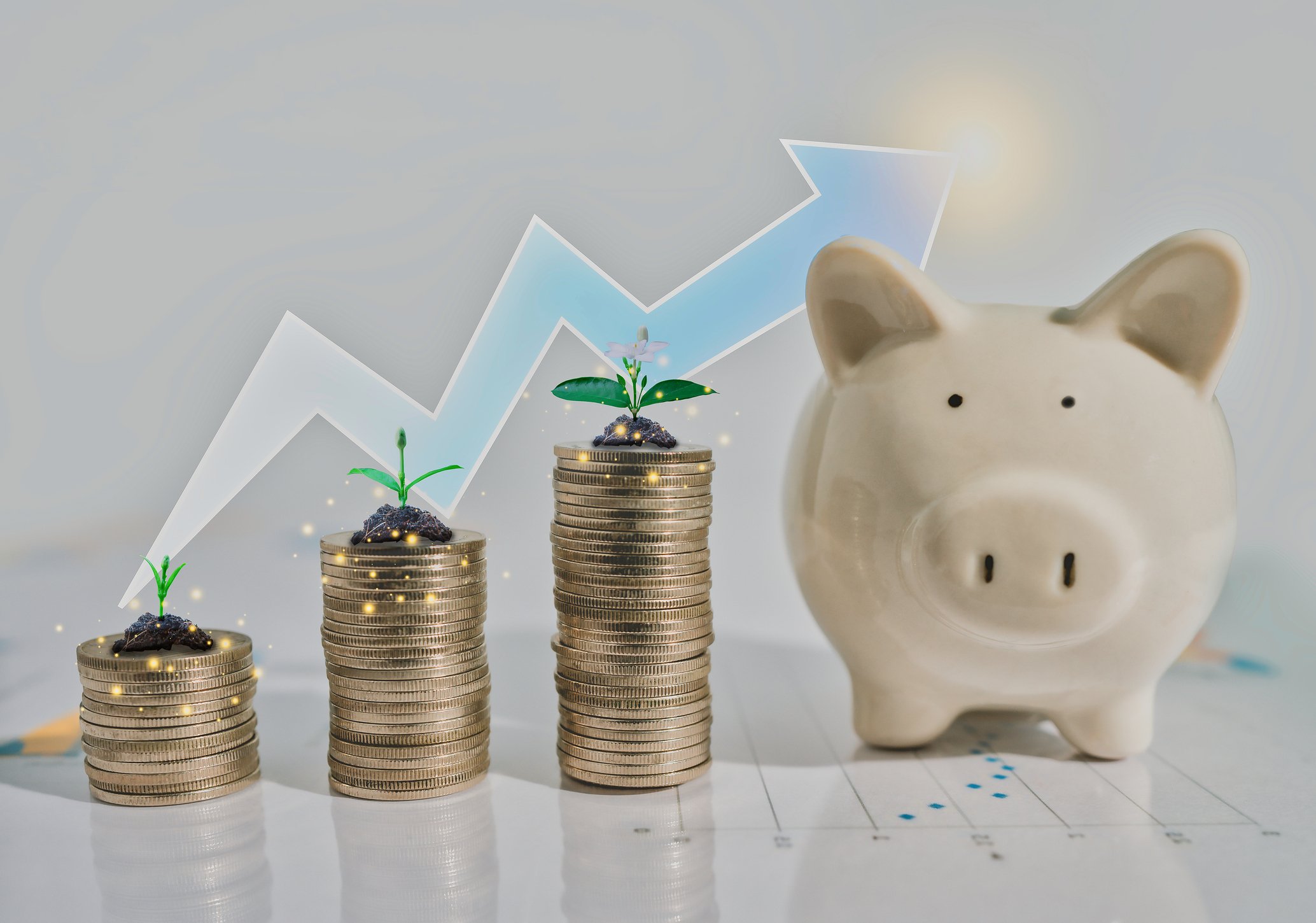 Progressively taller stacks of coins with sprouts on top on each, representing a rising stock chart, sit in front of an upward-trending arrow and next to a piggy bank.