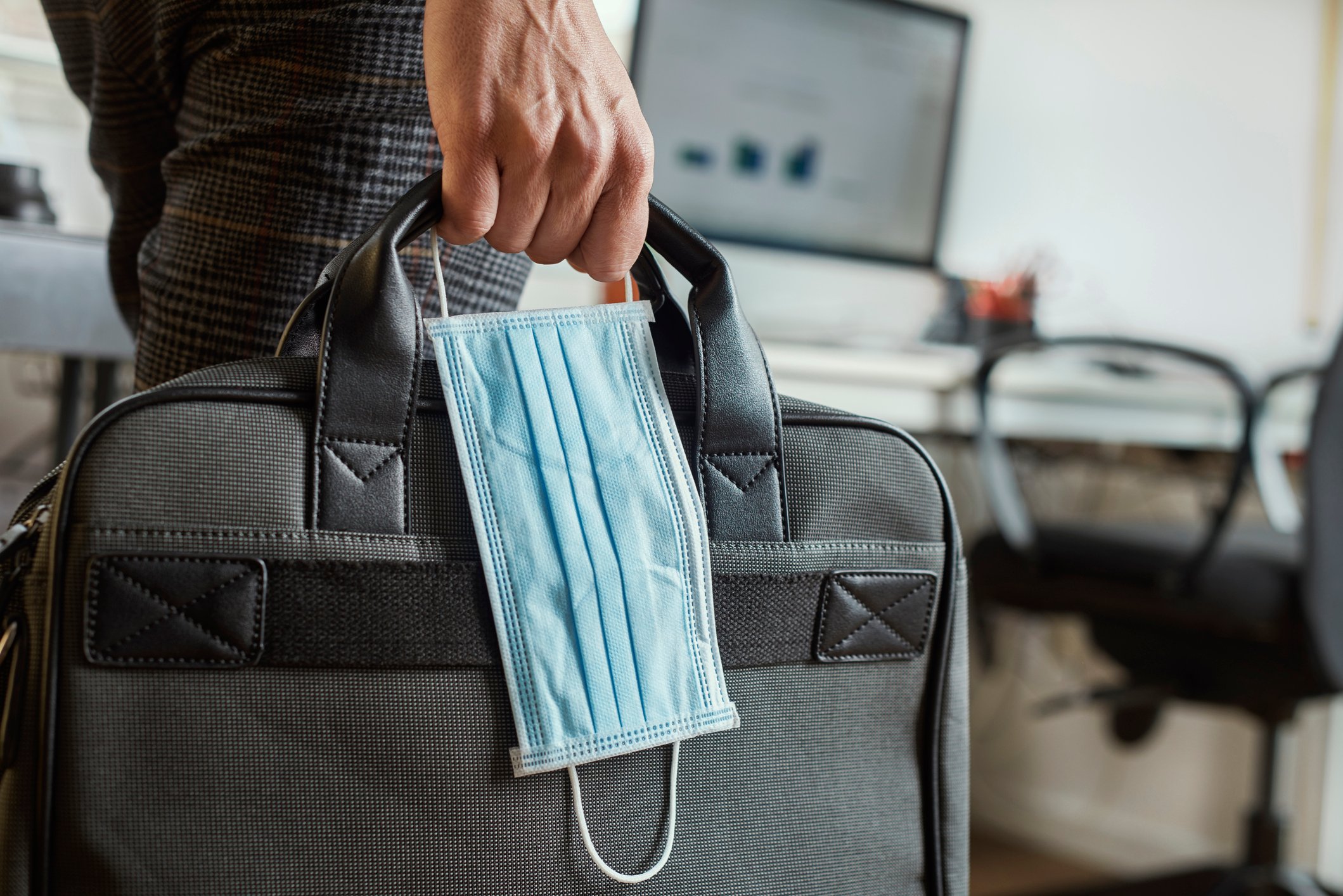 Closeup of a male hand holding a briefcase with a mask dangling from it. 