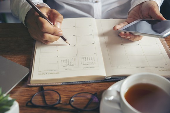 A guy holding his phone and writing information into a hard copy calendar book.