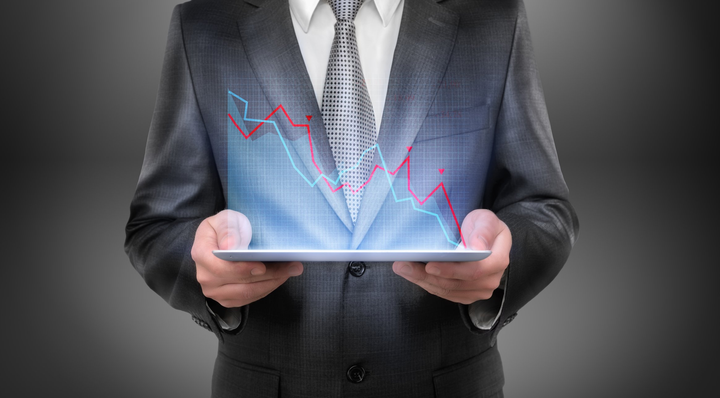 A person in a business suit holds a tablet displaying a downwardly sloping digital chart. 