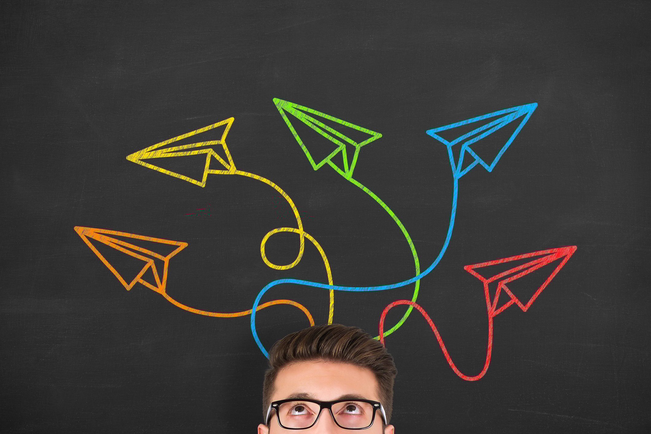 A young man glances up at a chalkboard above him, where colorful drawings of paper planes spiral out of his head.