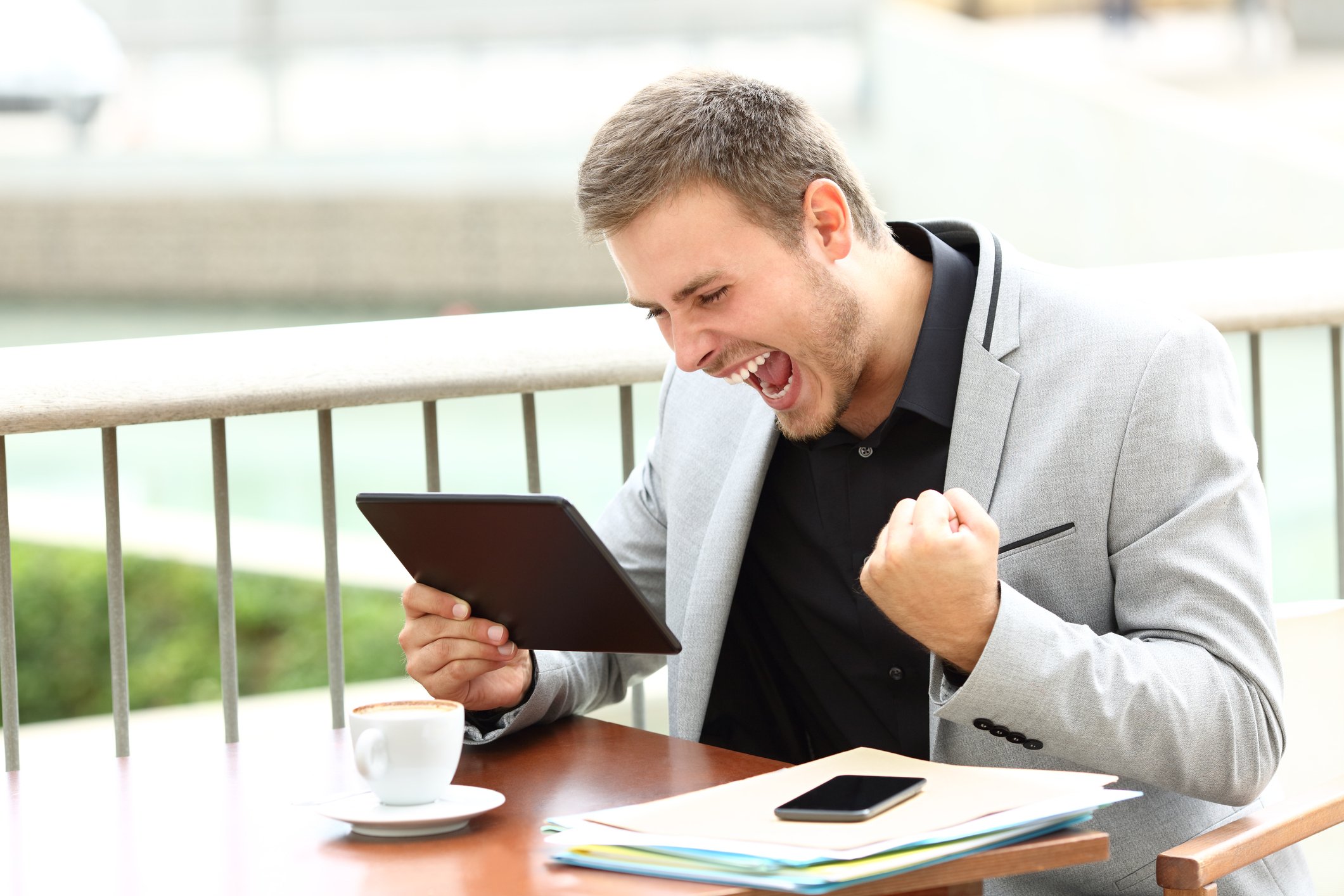 A smiling young businessman shouts and fist-pumps while looking at his tablet.