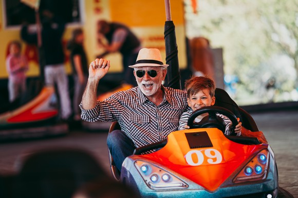 Older man in sunglasses and hat riding bumper car with young boy. 