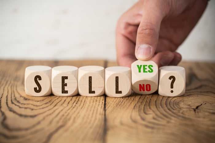 An investor's hand turns a block marked "yes" on one side and "no" on the other next to blocks spelling out "sell?"